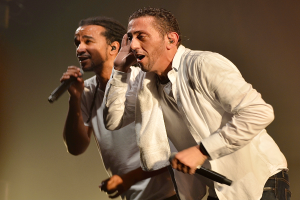 Zwei Männer in legerer Kleidung stehen nebeneinander auf einer Bühne, jeder hält ein Mikrofon in der Hand und scheint mitten in einer Performance zu sein, mit einem unscharfen Hintergrund.