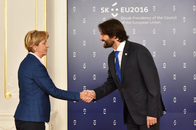 Bundeskanzlerin Angela Merkel und Bundeskanzler Joachim Gauck, die sich vor einer Wand mit einem Banner 'EU-Gipfel' und einer Tür im Hintergrund die Hände schütteln, beide lächeln.
