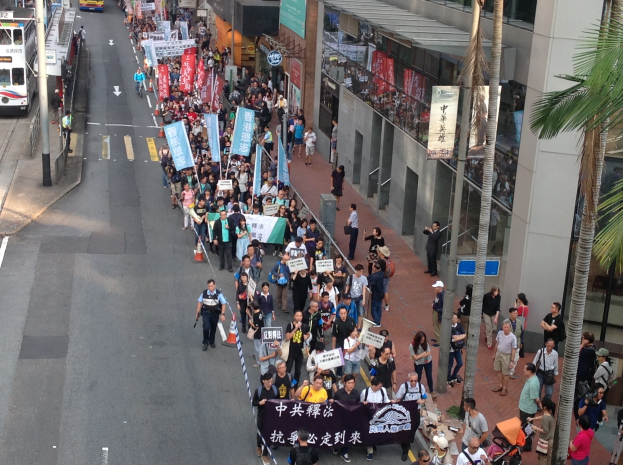 Eine große Gruppe von Menschen marschiert auf einer Stadtstraße und hält Schilder und Banner hoch, mit Bäumen, Gebäuden, Fahrzeugen und Straßenschildern im Hintergrund.
