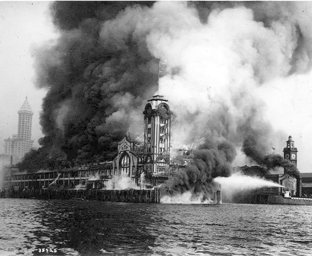 Schwarz-weiß-Foto eines großen, von Flammen umloderten Gebäudes mit Rauch, der herausquillt, einem Gewässer im Vordergrund und anderen Gebäuden und dem Himmel im Hintergrund.