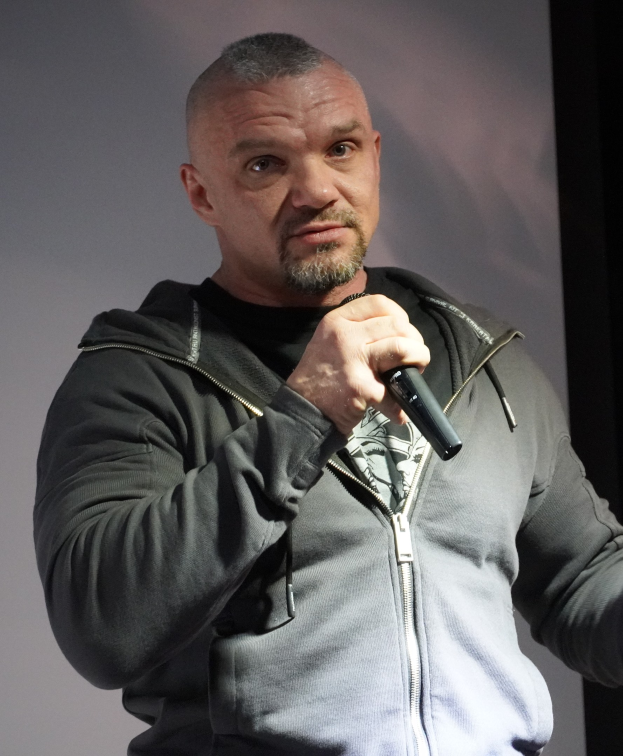 Comedian in a black suit and white shirt, holding a microphone, with slicked-back hair and a confident expression, standing in front of a wall.