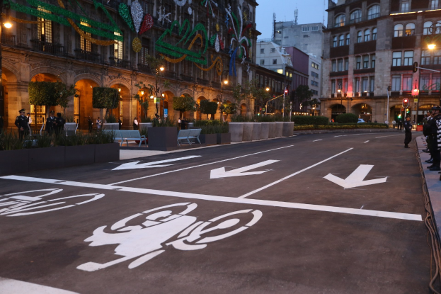 Stadtstraße mit einem markierten Fahrradweg, umgeben von Gebäuden, Straßenmöbeln, Fußgängern und Grünflächen unter einem klaren blauen Himmel.