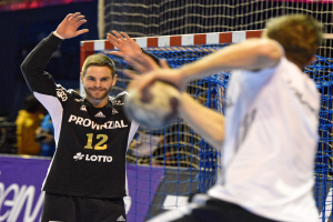 Zwei Männer spielen Handball vor einem Tor mit Netz und einer Banner mit Text im Hintergrund und einigen Zuschauern in einer Arena.
