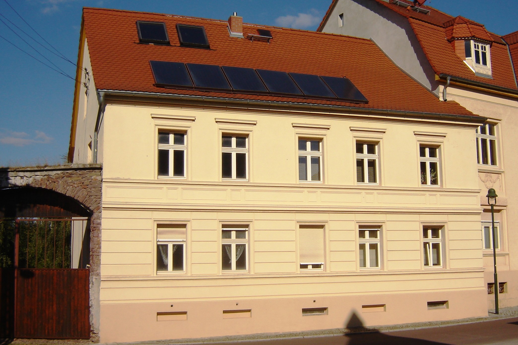 Gebäude mit Solarpaneelen auf dem Dach, umgeben von Straßeninfrastruktur einschließlich Pfosten, Laternen, Kabeln und Bäumen unter einem bewölkten Himmel.