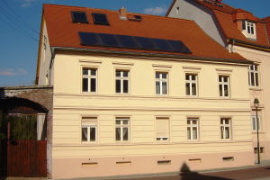 Gebäude mit Solarpaneelen auf dem Dach, umgeben von Straßeninfrastruktur einschließlich Pfosten, Laternen, Kabeln und Bäumen unter einem bewölkten Himmel.