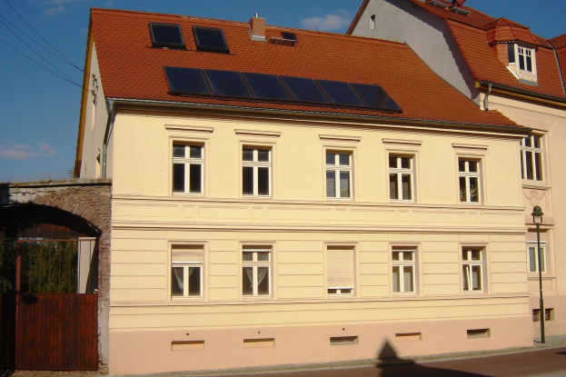 Gebäude mit Solarpaneelen auf dem Dach, umgeben von Straßeninfrastruktur einschließlich Pfosten, Laternen, Kabeln und Bäumen unter einem bewölkten Himmel.