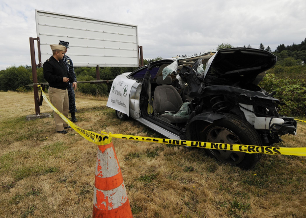 Ein Polizeibeamter in Uniform steht neben einem schwer beschädigten Auto am Straßenrand, mit eingedellter Motorhaube und zersplitterten Fenstern, während ein Verkehrskegel und ein Absperrband-marked Pfosten in der Nähe gegen einen Hintergrund von Bäumen und einem bewölkten Himmel sind.