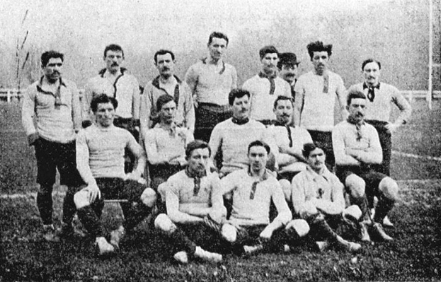 Schwarzes und weißes Foto einer Fußballmannschaft aus den 1920er Jahren, bei der einige Mitglieder stehen und andere auf dem Boden sitzen, vor Pfählen, Bäumen und einem klaren Himmel.