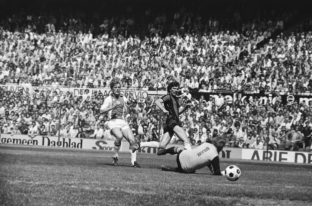Schwarz-weißes Foto von Männern, die auf einem Feld Fussball spielen, mit Zuschauern im Hintergrund und einer "Bundesliga 1978-79 Ajax 1-1 Hannover 96"-Tafel.