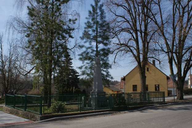 Ein Holocaust-Gedenkdenkmal steht in der Mitte einer Straße, umgeben von einem Metallzaun, umgeben von Gras, Bäumen, Häusern, einem Straßenpfahl und einem bewölkten Himmel.