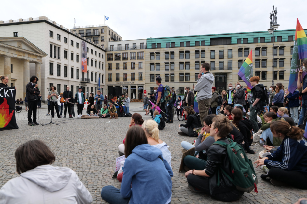 Eine Gruppe von Menschen, die auf dem Boden vor einer Menge sitzt, die Fahnen und Schilder hält, mit einer Person, die in ein Mikrofon spricht, einer Statue und Gebäuden im Hintergrund während einer anti-schwulen Demonstration in Berlin.