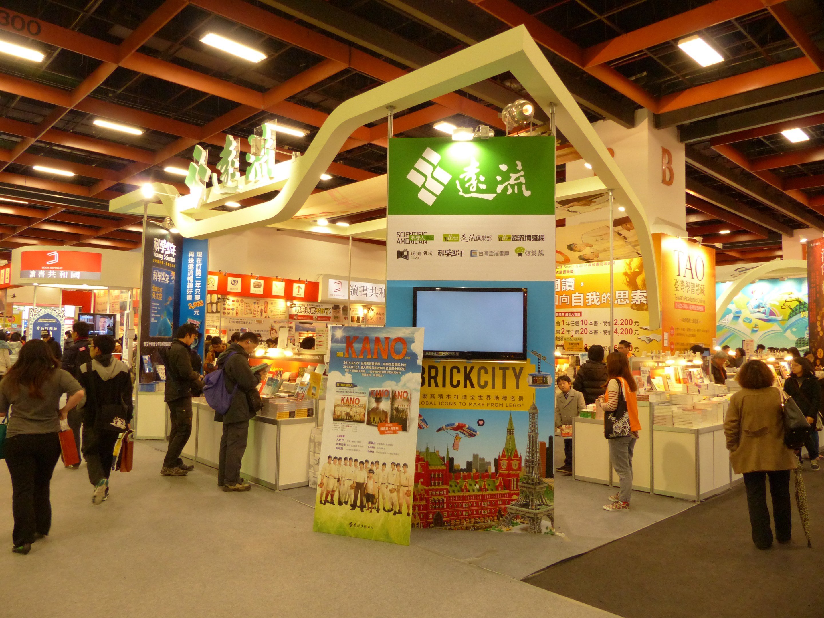 Menschen vor einem Messestand mit Bannern, einem Fernseher, Tischen mit Gegenständen, Schildern und Deckenleuchten.