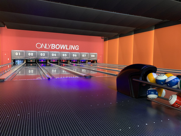 Eine Bowlingbahn mit Bowlingkugeln auf der rechten Seite, Bowlingkegel in der Mitte, einer textbedeckten Wand im Hintergrund und einer sichtbaren Decke.
