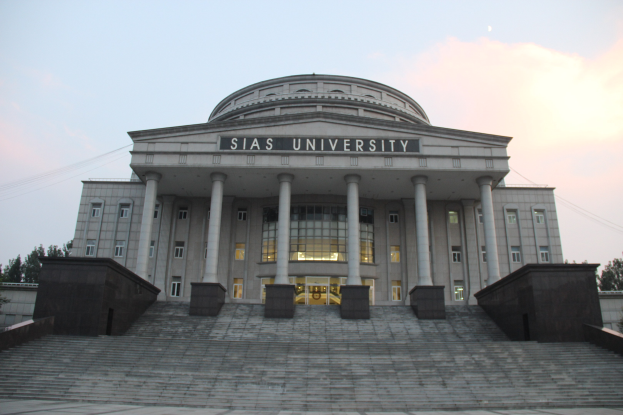 Großes Gebäude mit Säulen, Fenstern und Treppen, umgeben von Bäumen unter einem klaren blauen Himmel; der Name des Gebäudes ist auf der Fassade zu sehen.