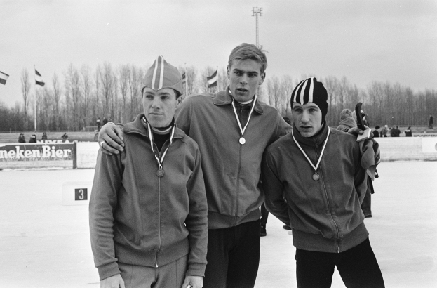 Drei Männer in Jacken und Medaillen stehen zusammen auf einem Eisstadion, mit einer Menge, Flaggen, Bäumen und einem Pfahl im Hintergrund und einer Tafel mit Schrift auf der linken Seite, alles in Schwarzweiß dargestellt.