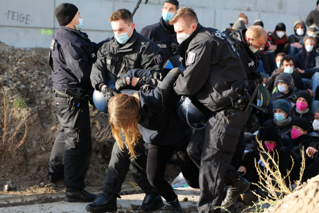 Gruppe maskierter Personen in Mützen, einige mit Waffen, umgeben eine Frau, die von der Polizei eskortiert wird, in einer städtischen Umgebung mit Pflanzen, Steinen und einer Wand mit Protesttext.