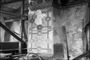 Ein Schwarz-Weiß-Foto eines alten, baufälligen Gebäudes mit einer gestalteten Wand, Holzpfählen und anderen Gegenständen, mit Text am unteren Rand.