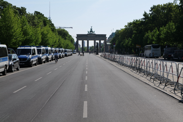 Lange Reihe von Polizeiwagen, die an der Straße vor dem Reichstagsgebäude in Berlin geparkt sind, mit Fahrradfahrern und Fußgängern, Barrieren, Bäumen, einem prunkvollen Bogen mit Statuen im Hintergrund und sichtbarem Himmel.