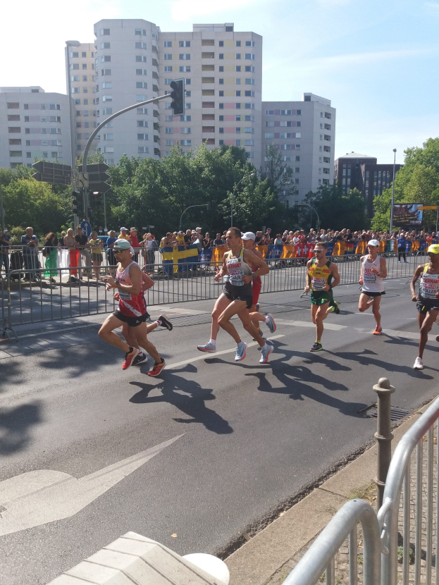 Eine Gruppe von Menschen, die bei einem Marathon auf einer Straße laufen, umgeben von einem Metallzaun, einem Schild, einem Straßenpfosten, einer Ampel, einigen Bäumen, einer Fahne mit Text, Gebäuden mit Fenstern und einem bewölkten Himmel.