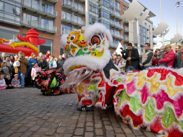 Ein lebendiges chinesisches Neujahrsfest in Amsterdam mit einem Löwen tanzen im Vordergrund, einer Menge Menschen mit Kameras und einem klaren blauen Himmel im Hintergrund.