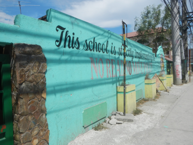 Grüne Wand mit Graffiti, das 'Diese Schule ist keine Stadt' liest, umgeben von Gebäuden, Bäumen, Strommasten, Kabeln und anderen städtischen Elementen unter einem bewölkten Himmel.