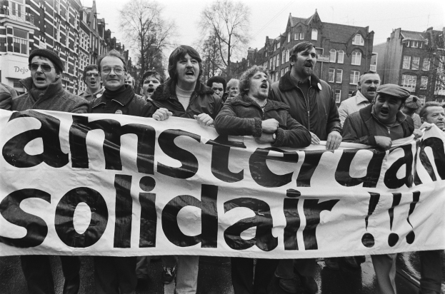 Eine Gruppe von Menschen mit einem Transparent mit der Aufschrift "Amsterdam Solidarität" auf einer Straße mit Bäumen, Gebäuden und einem klaren Himmel im Hintergrund.