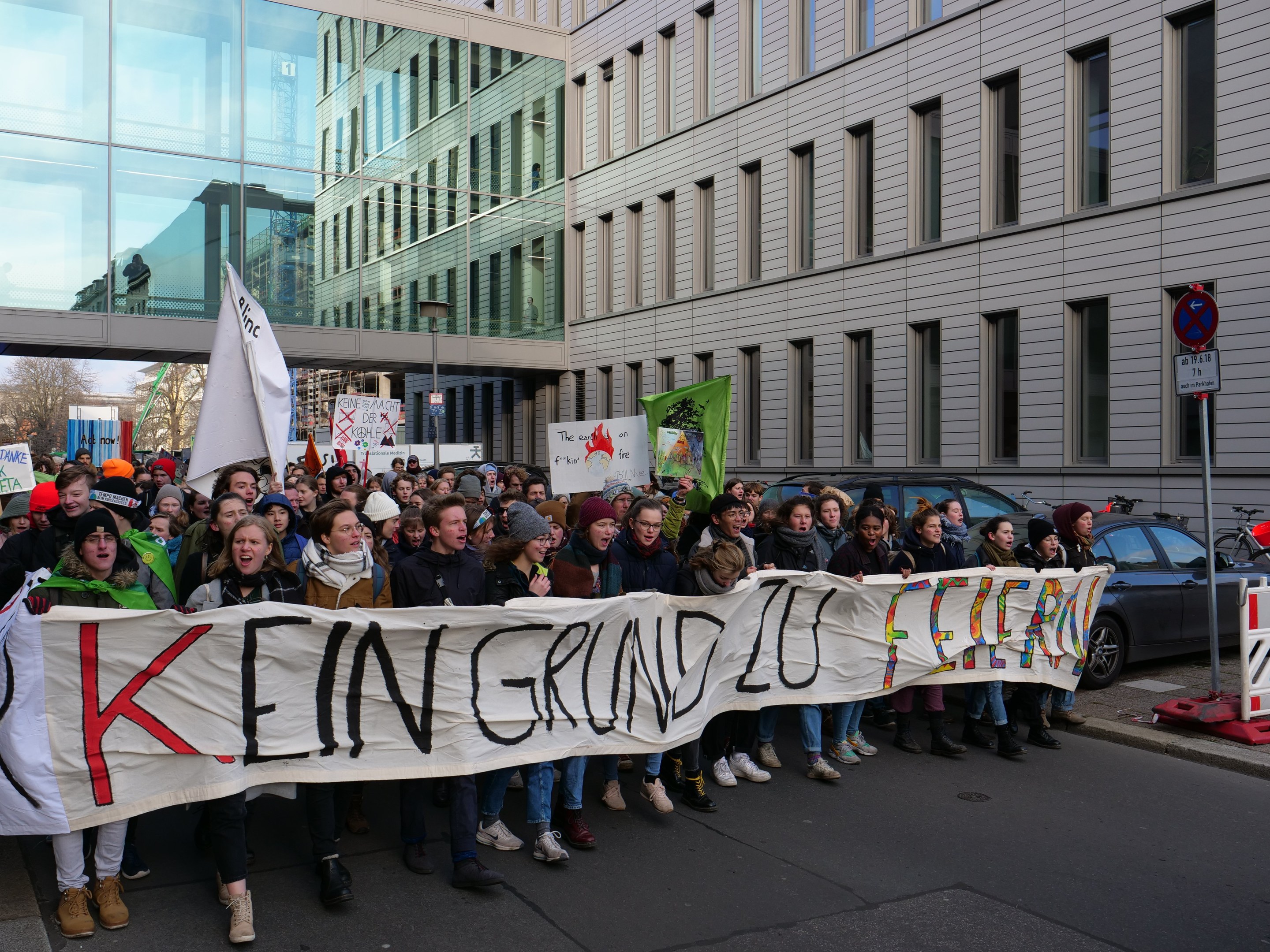 Protestierende mit Transparent 'Kein Grind Lo Feuer' gegen deutsche Sparpolitik, mit Gebäuden, Bäumen und Fahrzeugen im Hintergrund bei klarem blauem Himmel.