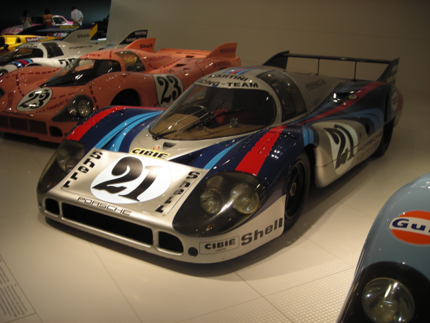 Ein Porsche 917K Rennwagen aus Le Mans in einem Museum neben anderen Sportwagen ausgestellt, mit Menschen in der Nähe und einer Wand im Hintergrund; ein Blatt mit Text ist links sichtbar.