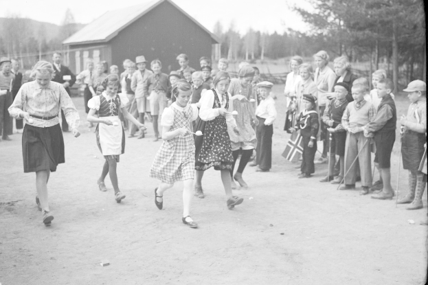 Schwarzes und weißes Foto von Kindern, die Fußball spielen, mit einer Menge von Zuschauern, die Fahnen halten, Bäumen und einem Haus im Hintergrund.