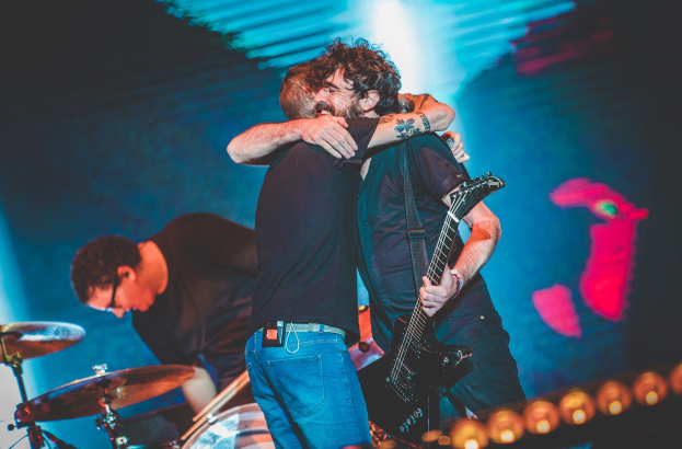 Zwei Männer umarmen sich auf der Bühne eines Konzerts, wobei einer eine Gitarre und ein anderer ein Schlagzeug spielt, beleuchtet von Scheinwerfern auf der Bühne.